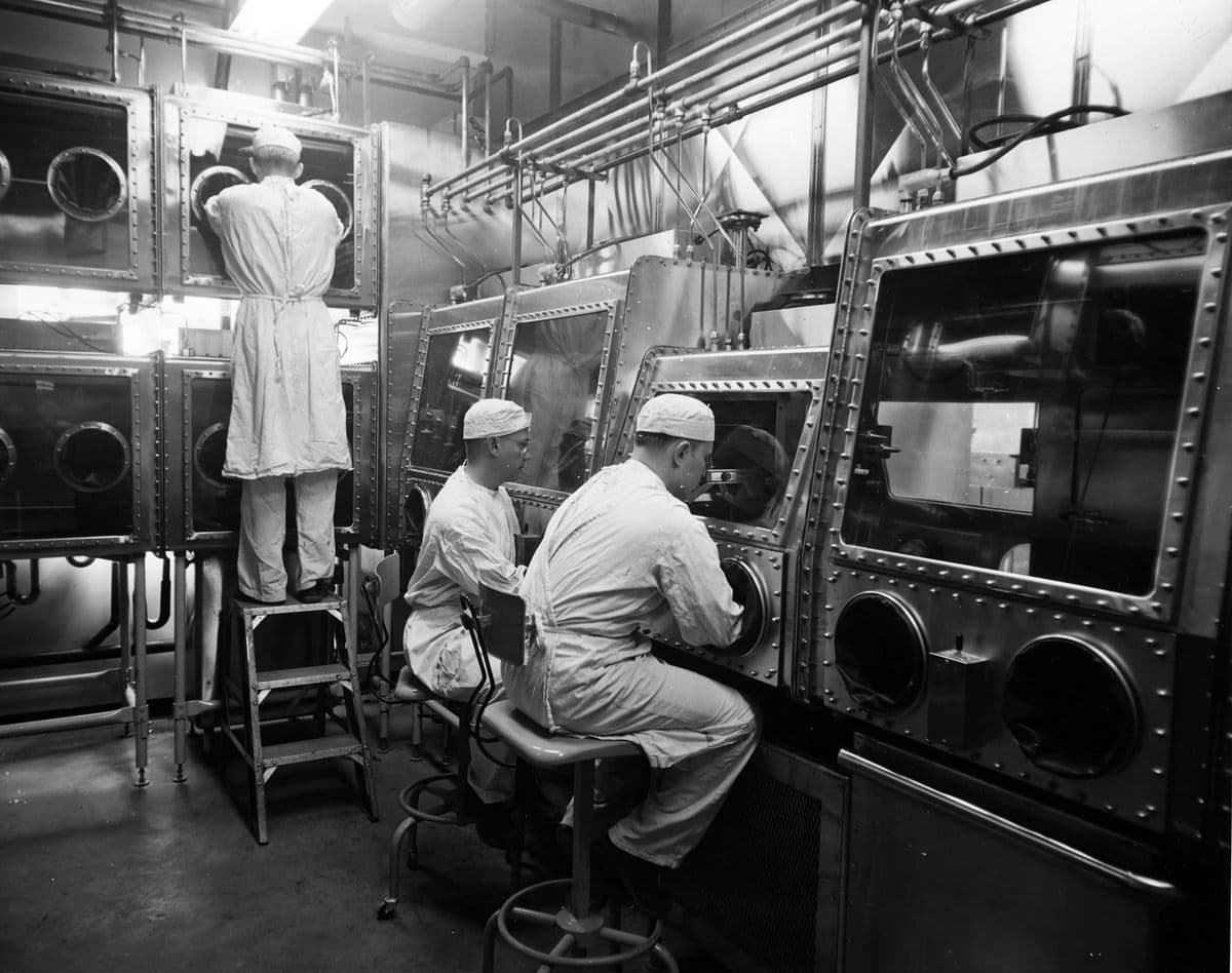 Resimde, laboratuvarda çalışan üç kişi görülüyor. Hepsi koruyucu giysiler giymiş ve yerleşik eldivenler ile kapalı bölmelerden malzemeleri taşıyorlar.